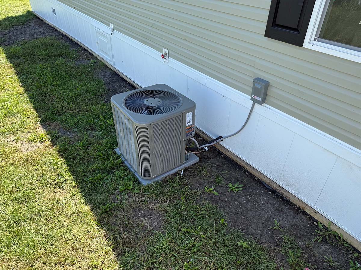 An outdoor air conditioning unit is installed next to the exterior wall of a light-colored building, sitting on a small concrete pad with grass and dirt surrounding it.