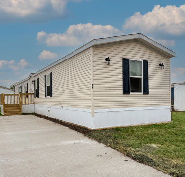 A beige mobile home with black shutters, a small wooden porch, and concrete driveway is pictured on a grassy lot under a partly cloudy sky.